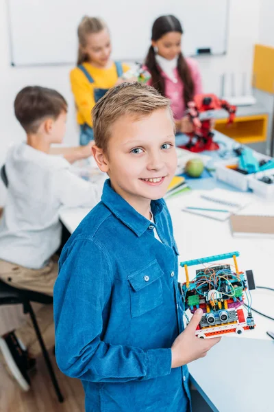 Sorridente scolaro che tiene robot e guarda la macchina fotografica con i compagni di classe sullo sfondo in classe — Foto stock