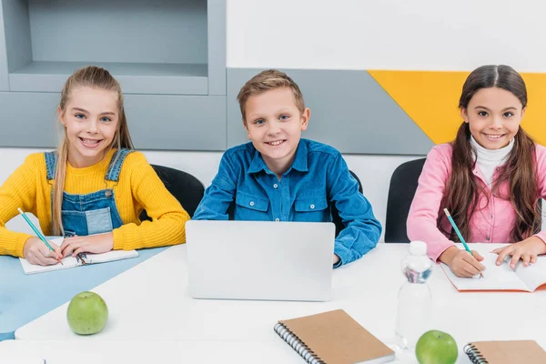 Des écoliers mignons écrivant dans des cahiers, utilisant un ordinateur portable et regardant la caméra en classe — Photo de stock