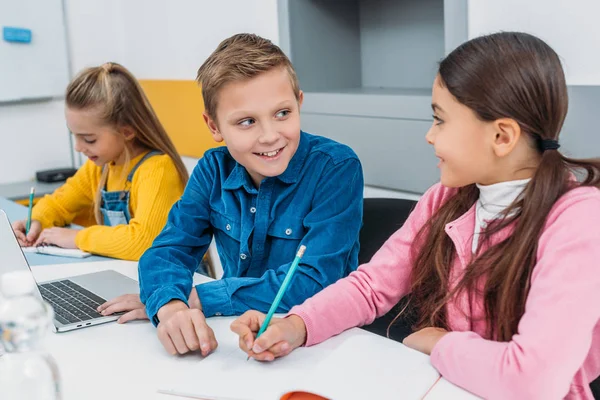 Crianças sorridentes sentadas na mesa e em sala de aula moderna — Fotografia de Stock