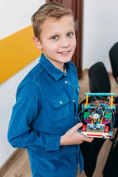 Écolier souriant debout, regardant la caméra et tenant robot fait main dans la classe de tige — Photo de stock