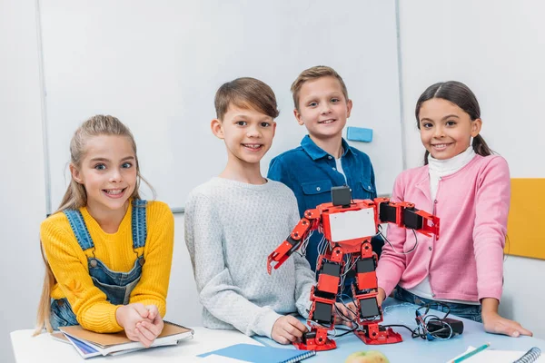 Heureux écoliers debout au bureau avec robot électrique rouge dans la classe de tige — Photo de stock