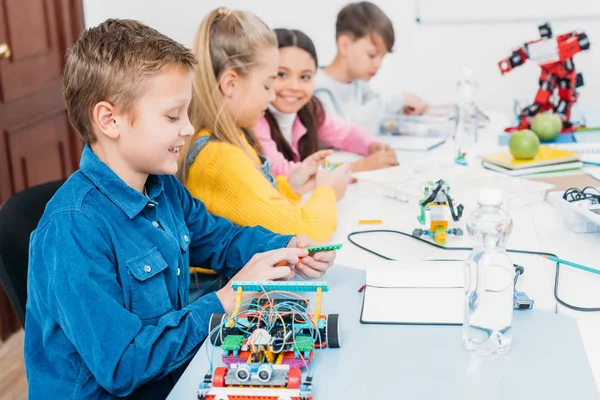 Niños felices sentados en el escritorio y haciendo robots en la clase de educación del tallo - foto de stock
