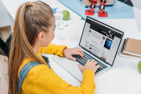 Colegiala utilizando el ordenador portátil con el sitio web de ciencia en la pantalla y artículo de lectura sobre robots - foto de stock