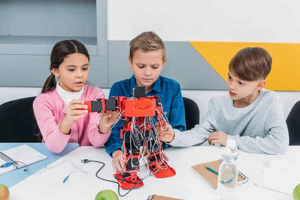 Escolares concentrados construyendo un gran robot eléctrico rojo en el escritorio durante la lección STEM - foto de stock