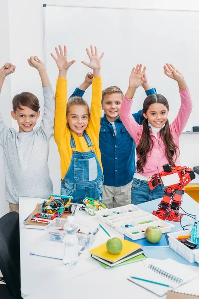 Escolares felices con manos levantadas de pie en el escritorio con robots de plástico y detalles en la clase de tallo - foto de stock