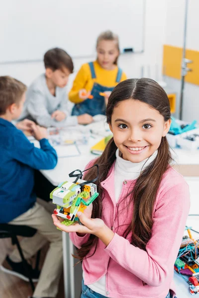 Écolière souriante tenant robot et regardant la caméra avec des camarades de classe en arrière-plan — Photo de stock