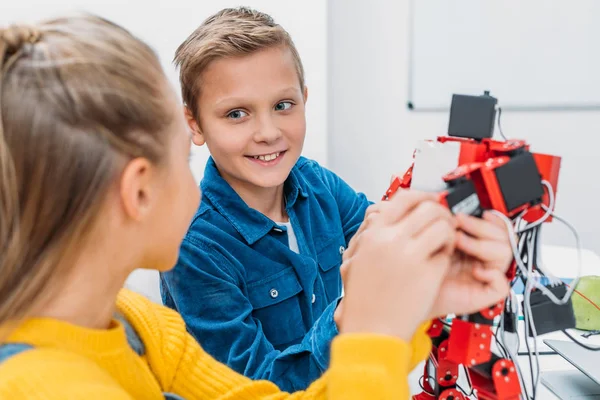 Schoolboy and schoolgirl looking at each other and touching handmade robot in stem education class — Stock Photo
