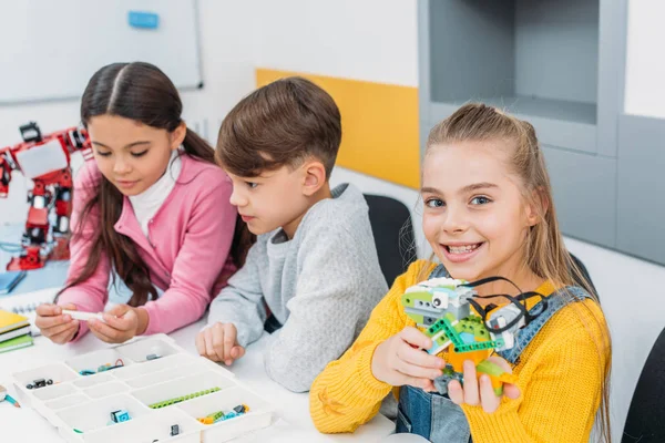 Lächelndes Schulmädchen hält bunten Roboter in der Hand, während Klassenkameraden Schachteln mit Details betrachten — Stockfoto