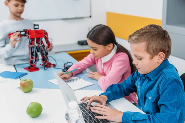 Children writing in notebooks, typing laptop and touching red robot in stem class — Stock Photo