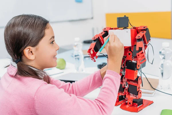 Smiling schoolgirl writing with pencil on red robot chest in stem class — Stock Photo