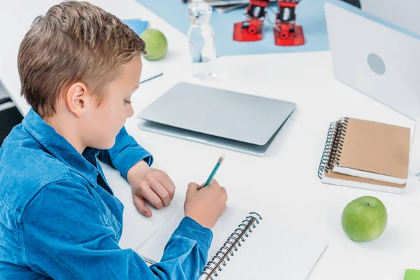 Colegial sentado en el escritorio y la escritura en el cuaderno en la clase de tallo - foto de stock