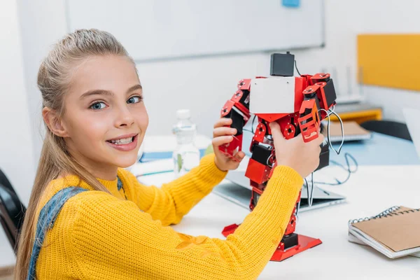 Smiling schoolgirl holding red electric robot and looking at camera in stem education class — Stock Photo