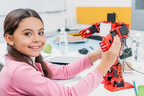 Entzückende Schülerin sitzt am Tisch, hält Robotermodell im Klassenzimmer und schaut in die Kamera — Stockfoto