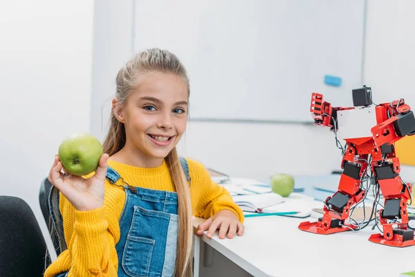 Colegiala sonriente sentada en la mesa con modelo robot en el aula STEM, sosteniendo manzana y mirando a la cámara - foto de stock