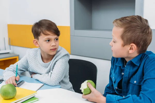 Scolaretti seduti alla scrivania, che si prendono una pausa, parlano e mangiano mele in classe — Foto stock