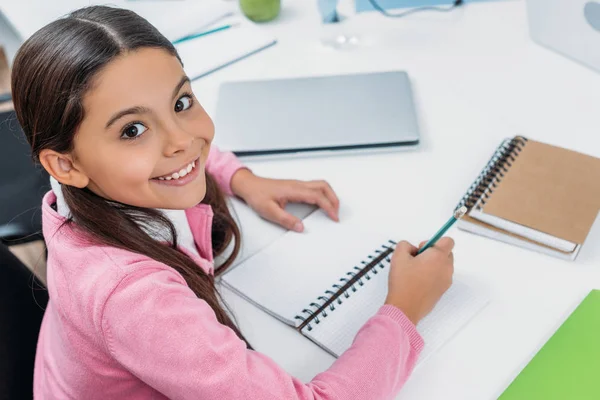 Entzückendes Schulmädchen schaut in die Kamera und schreibt während des Unterrichts in ein Notizbuch — Stockfoto