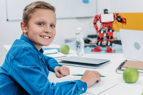 Écolier assis au bureau avec un modèle de robot, regardant la caméra et écrivant dans un cahier pendant la leçon de STIM — Photo de stock