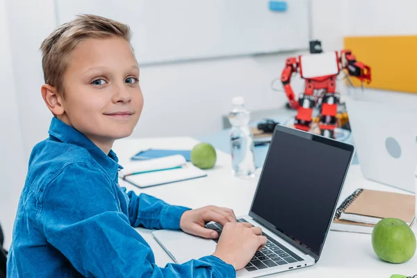 Colegial sentado en la mesa con el modelo de robot, mirando a la cámara y utilizando el ordenador portátil con pantalla en blanco durante la lección STEM - foto de stock