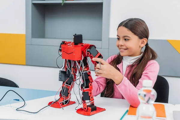 Adorable écolière assise à table, souriante et tenant un modèle de robot pendant la leçon de STIM — Photo de stock