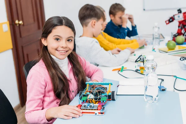 Adorable colegiala sosteniendo modelo robot, mirando a la cámara mientras sus compañeros de clase tienen lección STEM - foto de stock