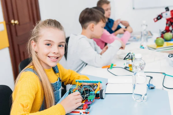 Adorable écolière tenant le modèle de robot, regardant la caméra tandis que les camarades de classe ayant leçon de STIM — Photo de stock
