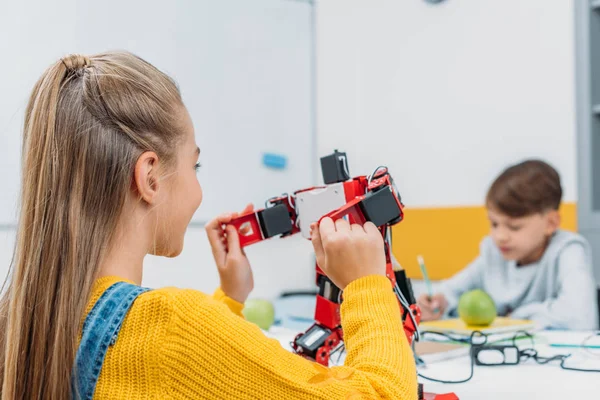 Schoolgirl sitting at table and holding robot model during STEM lesson with classmate on background — Stock Photo