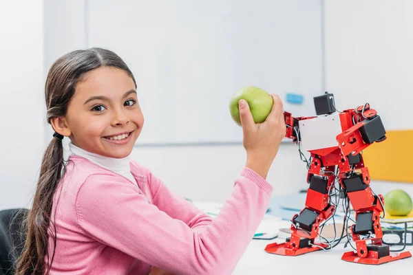 Adorable schoolgirl sitting at table with robot model at STEM classroom, holding apple and looking at camera — Stock Photo