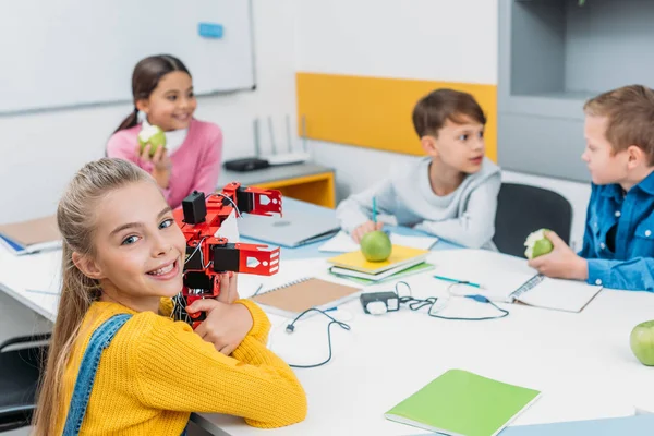 Happy schoolgirl holding robot model while classmates eating apples on STEM project in classrom — Stock Photo