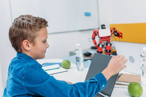 Scolaro seduto a tavola con modello robot e utilizzando il computer portatile durante la lezione STEM — Stock Photo