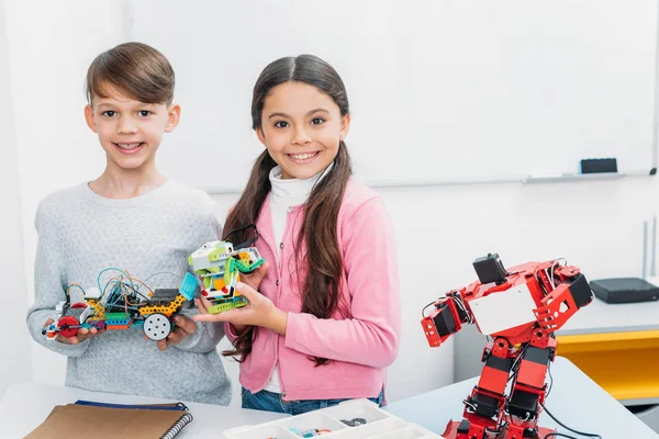 Joyeux écoliers regardant la caméra et présentant des modèles de robots faits à la main à la leçon de STIM — Photo de stock