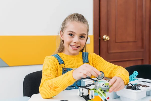 Fröhliches Schulmädchen blickt in die Kamera und arbeitet während der Vorlesung an einem handgefertigten Robotermodell — Stockfoto