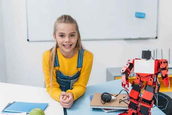 Entzückendes Schulmädchen lächelt, blickt in die Kamera und sitzt mit Robotermodell am Tisch im Klassenzimmer — Stockfoto