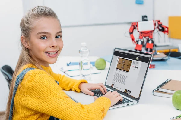 Colegiala sonriente sentado en la mesa con el modelo de robot, mirando a la cámara y el uso de la computadora portátil con el sitio web de ciencia en la pantalla durante la lección STEM - foto de stock