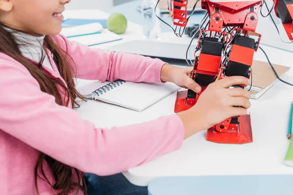 Vista parcial de la colegiala sentada en la mesa y jugando con el modelo de robot en la lección STEM - foto de stock