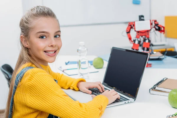 Colegiala sonriente sentada en la mesa con modelo robot, mirando a la cámara y utilizando el ordenador portátil con pantalla en blanco durante la lección STEM - foto de stock