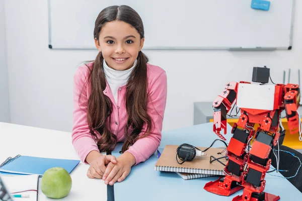 Adorable sonriente colegiala sentada en la mesa con modelo robot y mirando a la cámara en el aula STEM - foto de stock