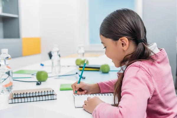 Schoolgirl writing in notebook during lesson in classroom — Stock Photo
