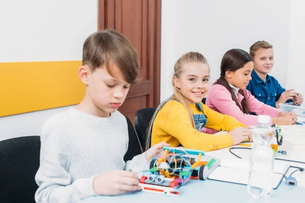 Adorable écolière regardant la caméra tandis que les camarades de classe travaillent au bureau pendant la leçon de programmation STIM — Photo de stock