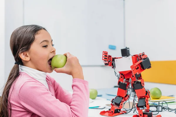 Schoolgirl eating apple and sitting at table with robot model at STEM classroom — Stock Photo