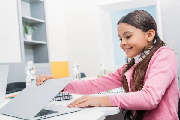 Colegiala sonriente sentada en el escritorio y apertura de la computadora portátil durante la lección en el aula - foto de stock