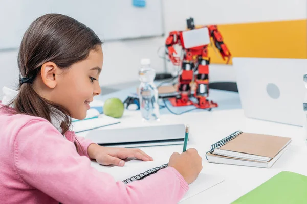 Scolaretta seduta alla scrivania con modello robot e scrittura su taccuino durante la lezione STEM — Foto stock