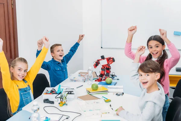 Niños felices animando y levantando las manos en el aire durante la lección STEM en clase - foto de stock