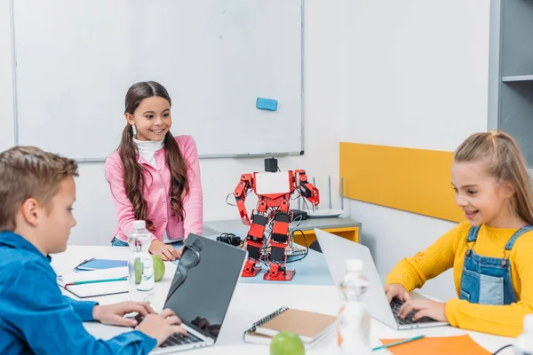 Sonrientes escolares que programan robots juntos y usan computadoras portátiles durante la clase educativa STEM - foto de stock