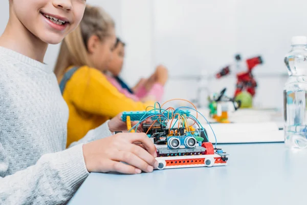 Vista recortada de colegial sosteniendo modelo robot hecho a mano y compañeros de clase trabajando juntos en el proyecto durante la lección STEM en segundo plano - foto de stock