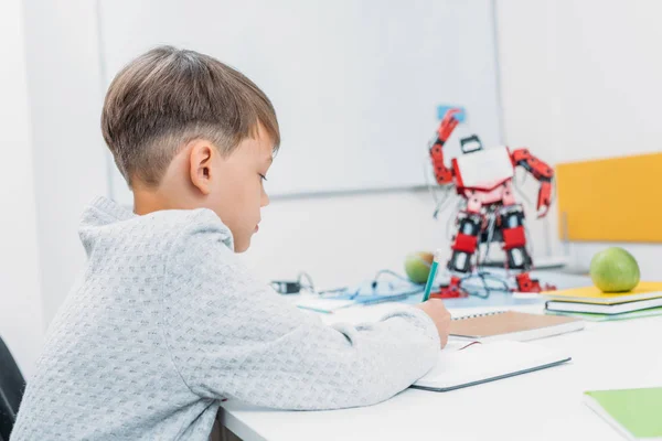 Colegial sentado en el escritorio con el modelo de robot y la escritura en el cuaderno durante la lección STEM — Stock Photo