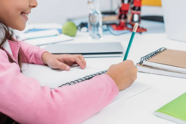 Vue recadrée de l'écolière au bureau écrivant dans un cahier pendant les cours en classe — Photo de stock