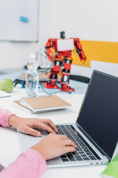 Cropped view of schoolgirl sitting at table with robot model and using laptop with blank screen during STEM lesson — Stock Photo