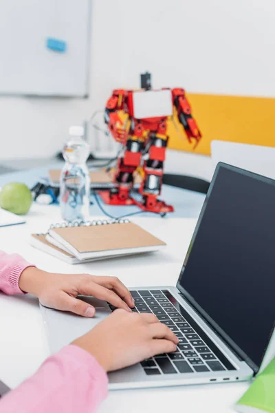 Vista recortada de la colegiala sentada en la mesa con el modelo de robot y el uso de ordenador portátil con pantalla en blanco durante la lección STEM - foto de stock