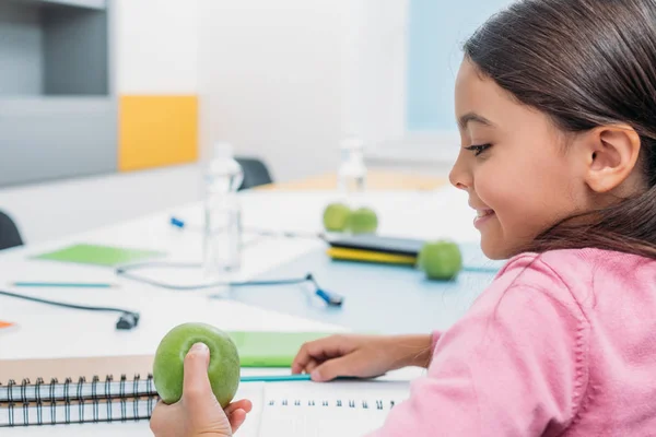 Entzückendes Schulmädchen schaut auf Apfel, während sie am Schreibtisch im Klassenzimmer sitzt — Stockfoto