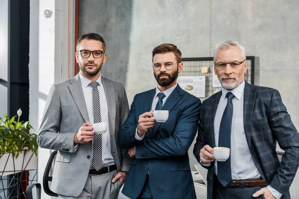 Three confident businessmen holding cups of coffee and looking at camera in office — Stock Photo